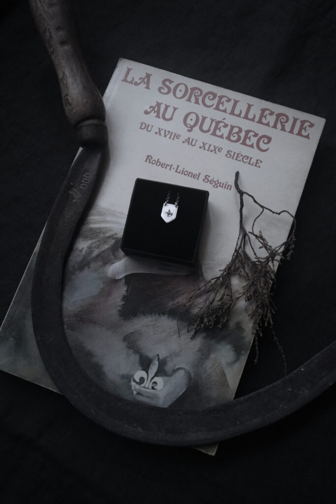 shield shaped pendant stamped with a fleur de lys stamp, with an old scythe and "Sorcellerie au Québec" book